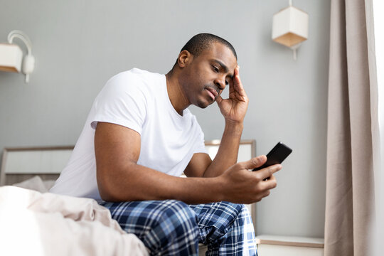 African american man sitting on bed looking at smartphone with worried expression in bedroom. Stressful lifestyle moment at home, communication, problem solving and anxiety concept
