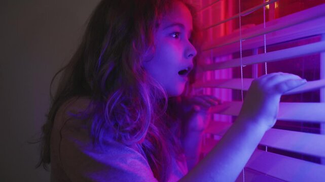 Afraid child hides alone in dark room. Afraid kid watches police flashing lights outside. Afraid child feels unsafe at night. Afraid kid portrait.