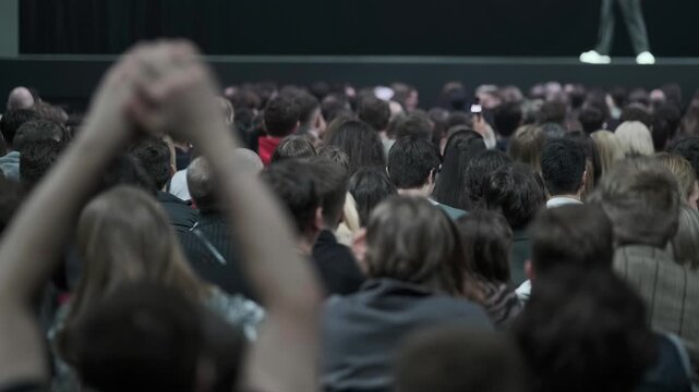 Large indoor audience applauding and raising hands, energetic crowd at live concert or conference, diverse people celebrating, excitement, unity, participation.