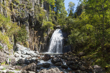 A secluded waterfall cascades over rugged cliffs into a rocky mountain stream, surrounded by lush green trees and sunlit birch, evoking serene wilderness, fresh water, and adventure. Waterfall Korbu © evannovostro