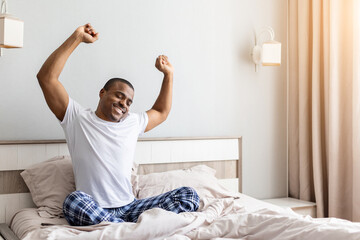 African american man stretching arms while sitting in bed in bedroom. Morning routine scene showing wake up, energy, freshness and healthy lifestyle at home © Anastasiya