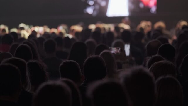 Large indoor audience watching bright screen at live event, silhouettes in dim light, some people recording with smartphones.