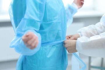 Naklejka premium Medical assistant putting isolation gown on doctor indoors, closeup