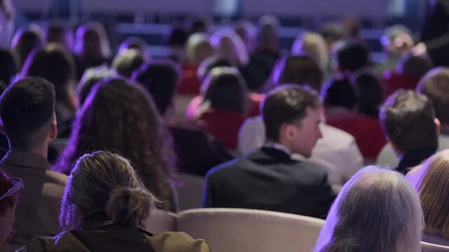 Audience seated in dim conference hall, listening to speaker, focused attention, professional gathering, networking potential, seminar atmosphere.