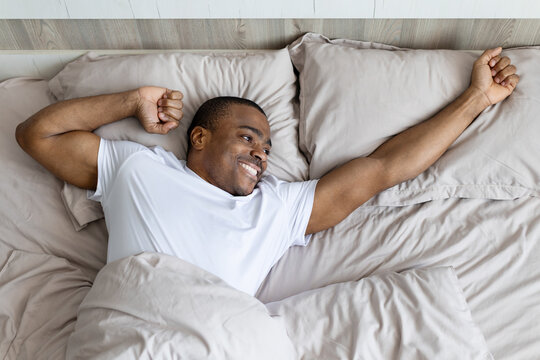 African american man stretching in bed after waking up smiling. Home lifestyle concept of morning routine, energy, relaxation and positive start of day