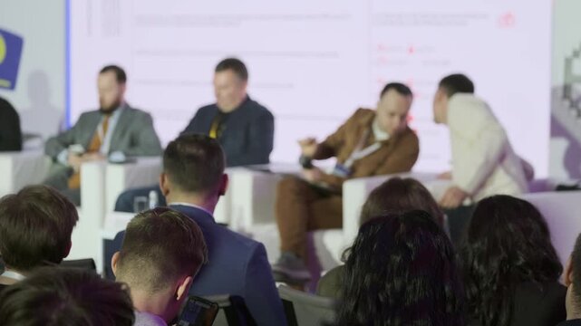 Business panel discussion at conference. Audience listening to speakers on stage, taking notes, using phones, and networking after session.