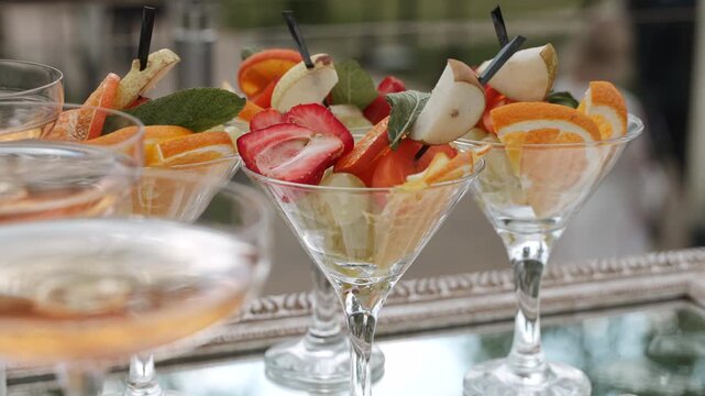 Compote dishes in form of glasses with cut fruits next to glasses with sparkling wine at buffet. Snack area at festive outdoor event. Presentation and treats for guests and influencers.