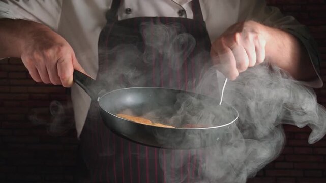 Chef in striped apron skillfully flips pancakes in a frying pan, with steam rising and a textured brick wall in the background