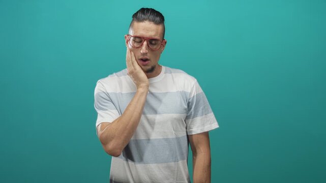 Man touching cheek with hand wearing red glasses and striped t shirt, goatee visible, standing against a teal studio backdrop with short sleeves; pain.
