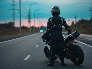 Naklejka premium motorcycle driver stands on the road by his motorcycle. Posing against the sunset . The concept of freedom and lifestyle of a biker