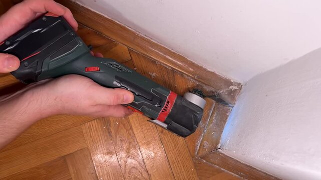 Hands use a cordless oscillating multitool to cut along a wooden baseboard corner, close indoor view shows home repair work and a focused practical mood