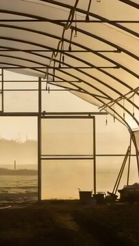 Vertical video: Framing arched ribs in hoophouse with sunlight warming doorway to fog, buckets