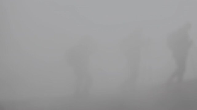 Silhouettes of group of people with backpacks and trekking poles walking through thick smoke or fog during group hike in mountains. Slow motion. Travel and tourism concept