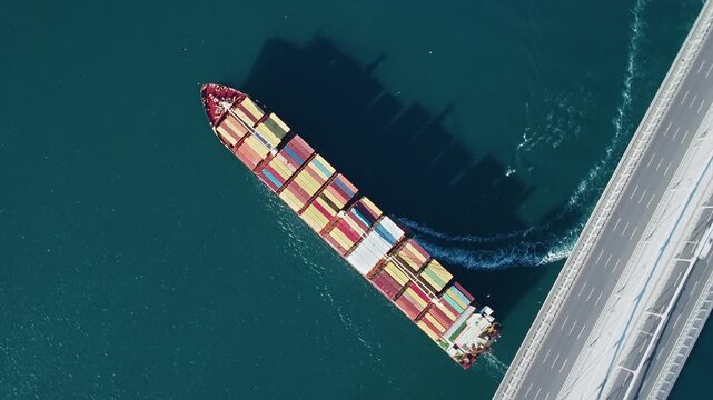 Top-down aerial view of container ship passing suspension bridge with an escort tug approaching starboard side, stern wave and water wake visible, Istanbul, Turkey