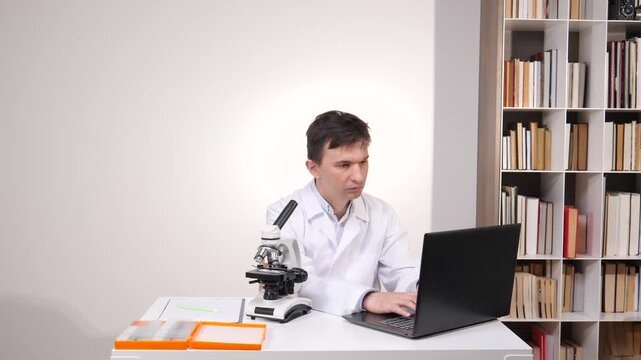 Male scientist in a white coat conducting research in a lab setting, using a microscope and laptop while surrounded by bookshelves, symbolizing continuous learning and scientific discovery