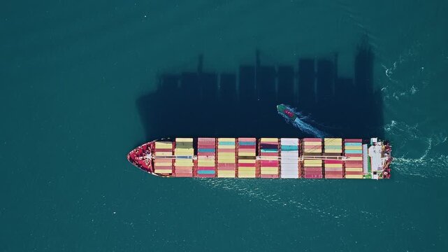 A container ship navigating the open waters as escort tugboat departs, colorful stacked containers and bridge shadow cast across deck. Top-down aerial view. Istanbul, Turkey