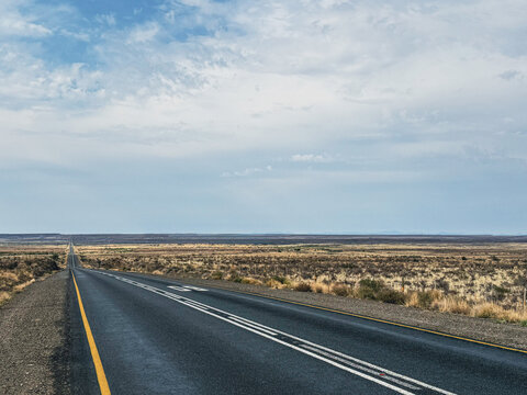 View of a long straight asphalt road stretching through the vast semi-arid landscape of the Karoo under a blue sky with scattered clouds in Beaufort West, Western Cape, South Africa.