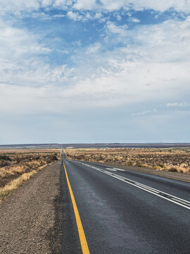 View of a long straight asphalt road stretching through the semi-arid landscape of the Karoo under a blue sky with scattered clouds Beaufort West, Western Cape, South Africa.