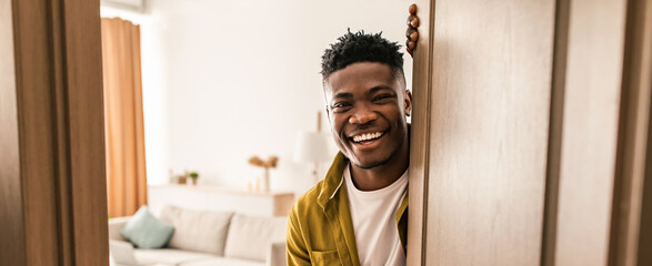 A man stands at the doorway with a smile. He has short hair and is wearing a green shirt. A bright living room can be seen behind him with a sofa and decorative items. © Prostock-studio