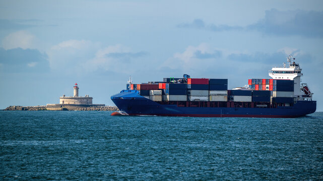 View of a large container ship with colorful cargo containers sailing past the Bugio Lighthouse under a cloudy sky in Lisbon, Lisbon, Portugal.