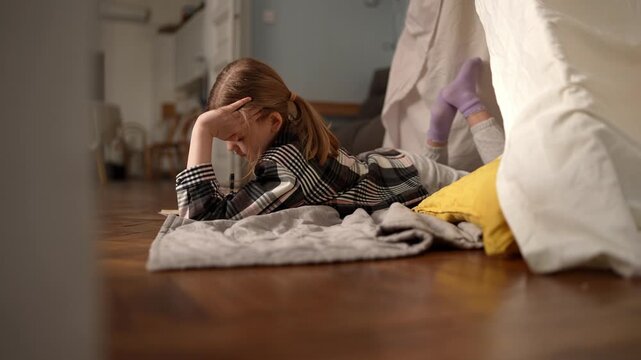 White girl focused on homework, lying on hardwood floor under blanket fort, pencil moving across notebook, determined posture, cozy pillow and yellow cushion, .