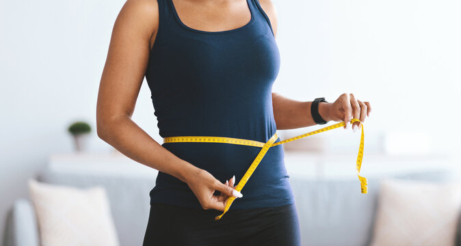 A woman stands indoors and uses a yellow measuring tape to measure her waist. She focuses on her body measurements as part of her fitness routine. The setting is bright and open.