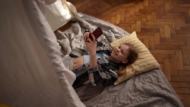 Young caucasian girl lying on pillow scrolling phone, overhead angle shows soft blanket and parquet floor, warm lamp glow, calm expression, evening solitude .