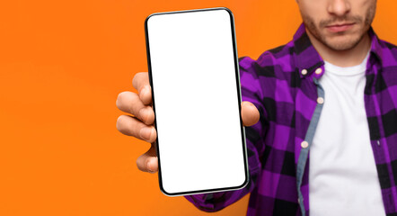 A man stands in front of a bright orange background and holds a smartphone with a blank screen. He is wearing a purple and black checkered shirt and a white t-shirt underneath. © Prostock-studio