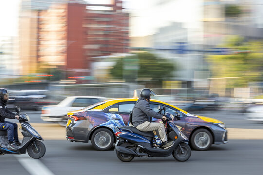 View of scooters with riders and cars rushing through a busy city intersection with motion blur and urban buildings in the background Taipei City, Taiwan.
