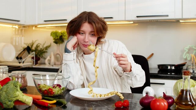 Young woman looks at measuring tape with sad face while trying healthy diet meal in home kitchen. Adult girl feels stress from weight loss goal and tests willpower during clean eating salad routine.