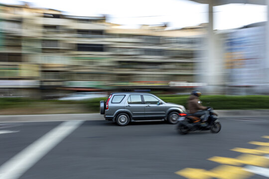 View of a grey SUV and a scooter rider moving fast through a city street with motion blur and urban buildings in the background Taipei City, Taiwan.