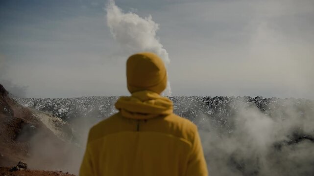 Man in yellow jacket and yellow knitted hat stands on edge of crater with solidified lava and watching as column of steam rises from cracks. Back view