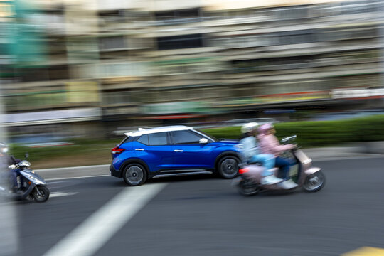 View of a blue SUV and scooters driving through a busy urban intersection with motion blur and city buildings in the background Taipei City, Taiwan.