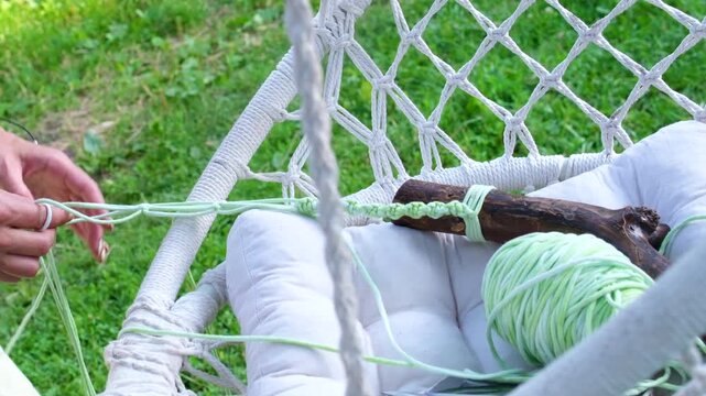 Female hands crafting a macrame piece from soft pastel green yarn outdoors, focusing on detailed knotting technique, handmade textile art in a calm summer garden atmosphere.