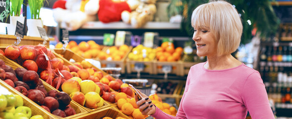 A woman stands in a grocery store looking at a variety of fruits. She holds a phone in her hand while choosing apples and pears. The store is colorful and busy with fresh produce. © Prostock-studio