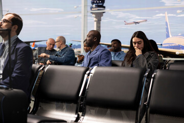 Fototapeta premium Men and woman in diverse crowd waiting in busy airport lounge, sitting on chairs bench. Group of multiethnic passengers enduring long flight layover before next departure