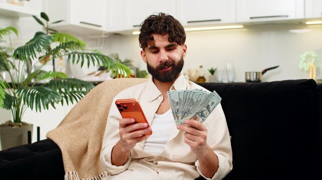 Indian man at home uses smartphone calculator counts dollar cash salary plans budget bills savings. Arabian guy checks orange mobile phone adds cost of rent feels calm focused careful about money.