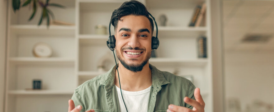 A man with headphones engages in conversation while making hand gestures. He sits in a well-lit room with shelves filled with various items. His expression shows enthusiasm and focus.