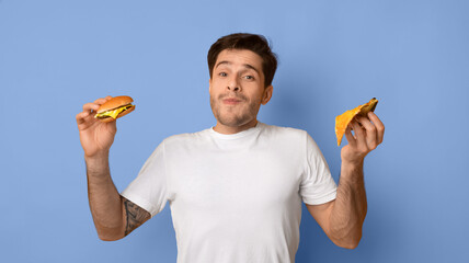A man stands in front of a light blue background holding a burger in his left hand and a taco in his right hand. He has a relaxed expression as he looks at the camera, enjoying the moment. © Prostock-studio