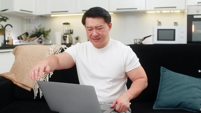 Asian man at home slams laptop shut after long work feels tired and raises hand for enough then sighs once. Chinese guy on sofa waves no rests back closes eyes a moment breathes out and feels relief