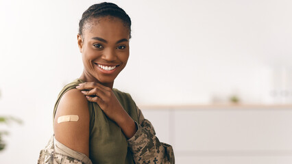 A woman smiles proudly while pointing to a bandage on her arm. She is dressed in a military uniform and stands in a bright clinic. This moment highlights her health initiative. © Prostock-studio