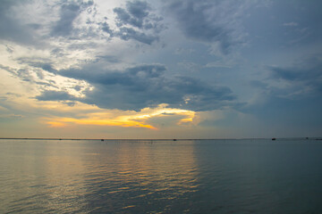 Obraz premium Golden sunset light breaks through layered storm clouds over a tranquil sea, creating soft reflections and a moody horizon with distant silhouettes.