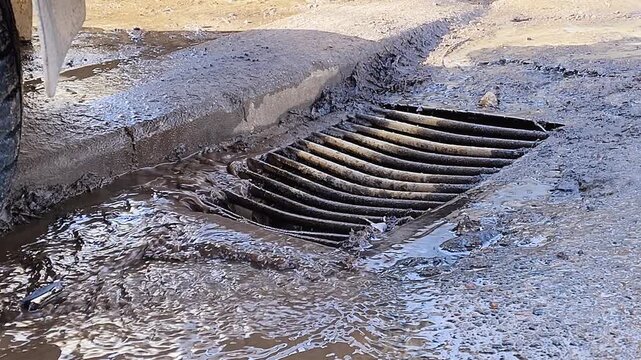 Dirty rainwater and mud flowing down the street into a storm sewer drain after heavy rain, representing issues like flooding, poor infrastructure, and environmental pollution from runoff