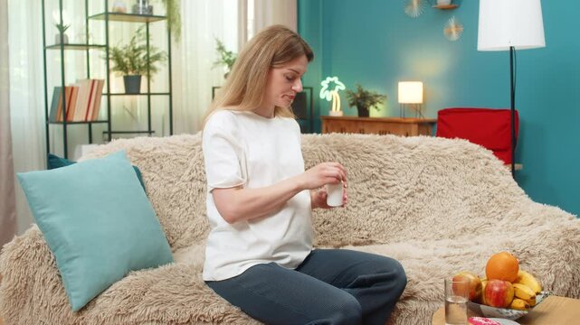 Pregnant middle-aged woman at home takes small pill with water feels calm and safe about vitamin care. Caucasian girl on sofa exhales relief believing daily supplement keeps baby strong and mom well.