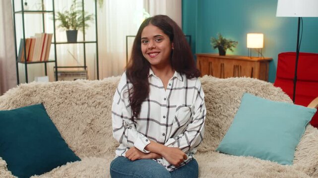 Indian woman at home forms heart with hands and blows kiss toward camera showing love affection greeting. Arabian girl on sofa smiles playfully sending romantic air kiss warm thanks gesture to viewer.