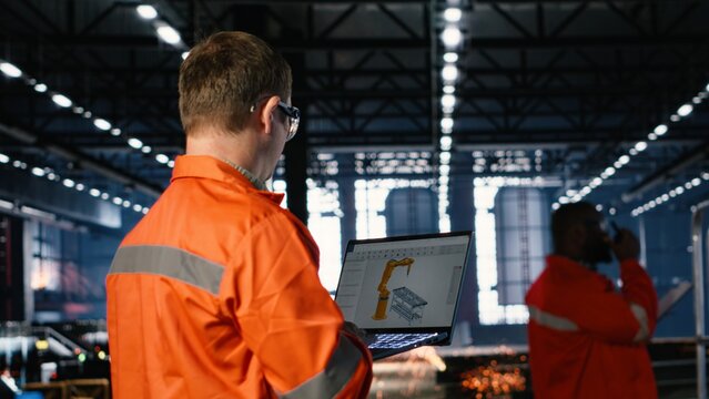 Staff uses advanced software on laptop to manage robot technology and production tools. Surrounded by industrial machinery and metal equipment, employee supports logistic operation.