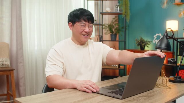 Asian man sitting at home table removes eyeglasses, leans back satisfied, closes laptop signaling end of final tasks. Chinese guy smiles quietly, enjoying sense of completion after productive session.