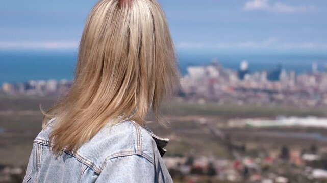 Blonde woman wearing a denim jacket enjoying the beautiful panoramic view of a coastal city with modern buildings and the sea, her hair blowing gently in the wind on a sunny day