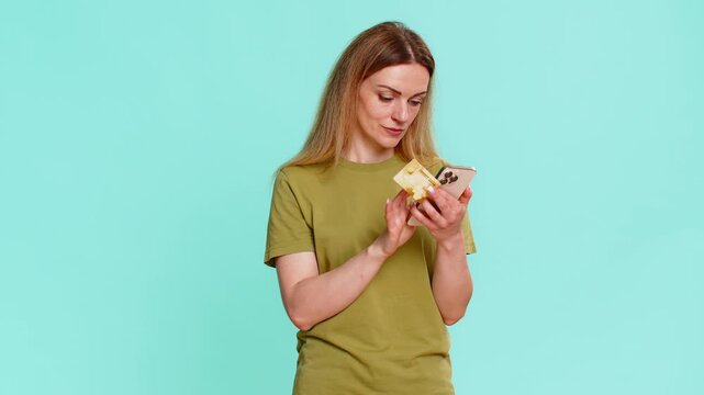 Middle-aged woman clicks order button while holding smartphone and credit card, showing digital payment moment with confidence. Adult girl on blue background, buying goods online with focused emotion.