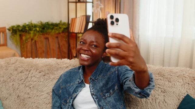 African American happy woman smiles joyfully while taking selfies on smartphone, trying different angles, expressions. Black girl enjoys the playful process of capturing trendy shots for social media.